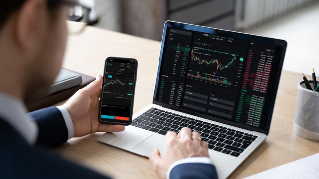 Trader watching charts on a laptop and phone at a desk